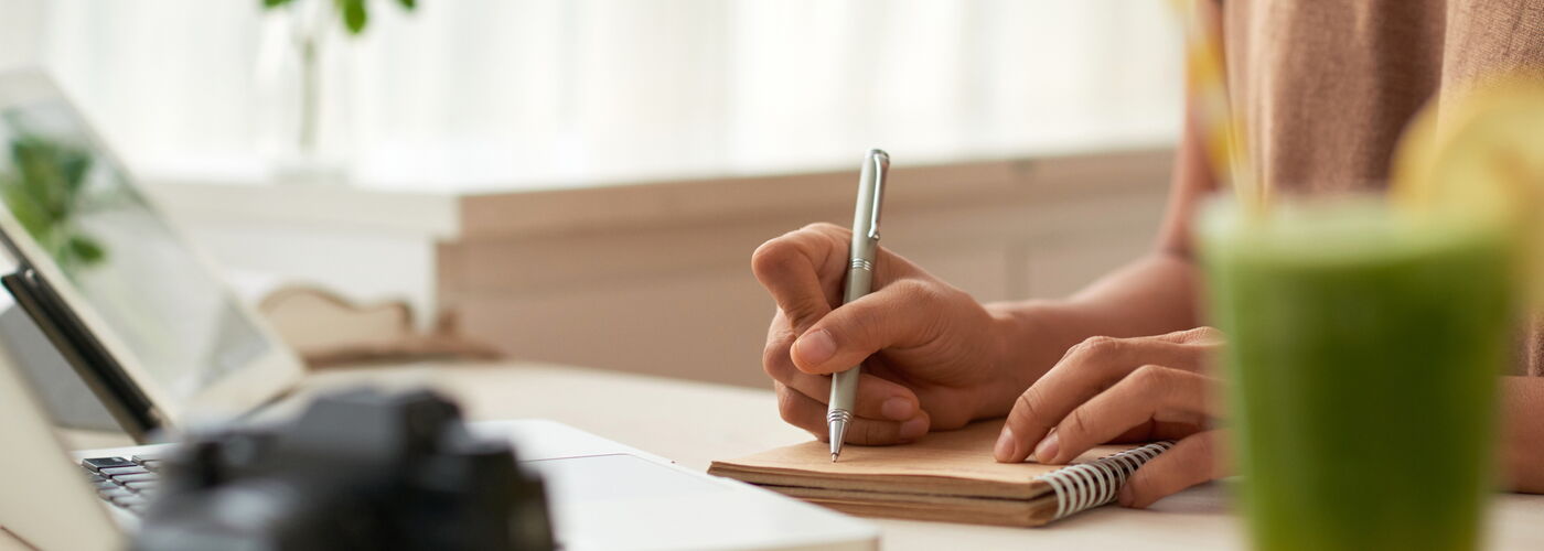 Eine Frau notiert etwas in einen Ringblock, vor ihr steht ein grüner Smoothie